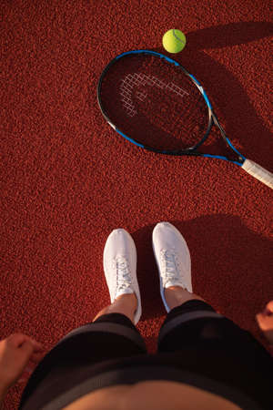 Sports woman athlete getting on outdoor red hard court in summer. Sporting event concept. Professional player preparing for tournament. Top view of feet in white sneakers and a tennis racketの写真素材