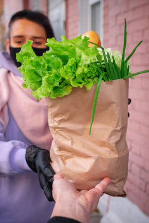 A girl in a black mask delivers a paper bag of groceries to a man customer. Ordering groceries from...