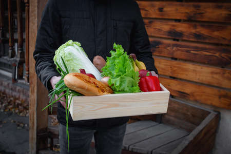 A man courier in a black jacket is holding a wooden box of groceries. Ordering groceries from an online store. Home delivery of groceries during a virus outbreak, virus panic, and pandemic.の写真素材