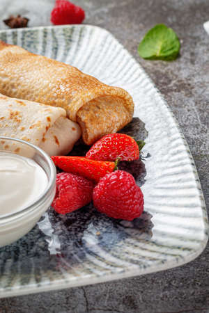 A delicious breakfast. Sweet fried pancakes filled with strawberries in a white plate with sour cream on a gray stone table with a napkin and cutleryの写真素材