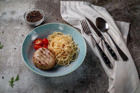 Spaghetti pasta with meat patty and fresh tomatoes on a blue plate against a gray stone table.の写真素材