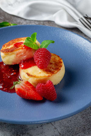 Fried pancakes with strawberry jam and raspberries and mint in a blue plate against a gray stone table. A sweet breakfast in the morningの写真素材