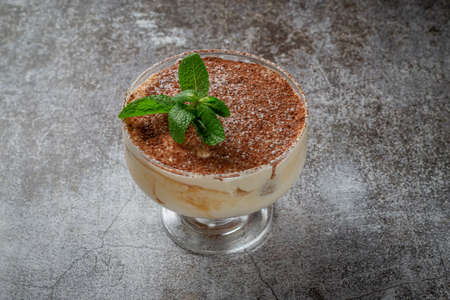 Classic tiramisu dessert with mint leaves in a glass, with cocoa and chocolate in the background of...