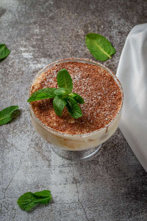 Classic tiramisu dessert with mint leaves in a glass, with cocoa and chocolate in the background of...