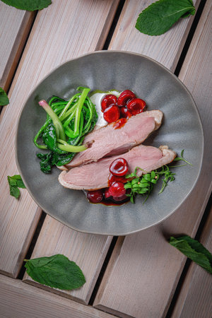 Tender duck meat and pickled cherries with herbs in a brown clay plate on a wooden tableの写真素材