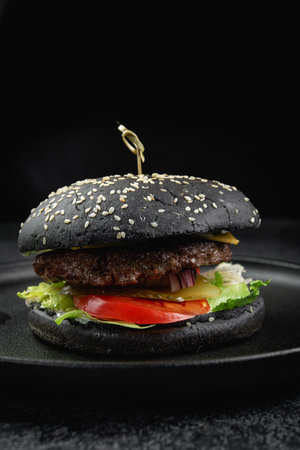 Black burger with a juicy meat patty, cheese, tomato, pickle on a dark stone tableの写真素材