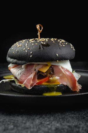 Black burger with bacon, cheese sauce, tomato on a dark stone tableの写真素材