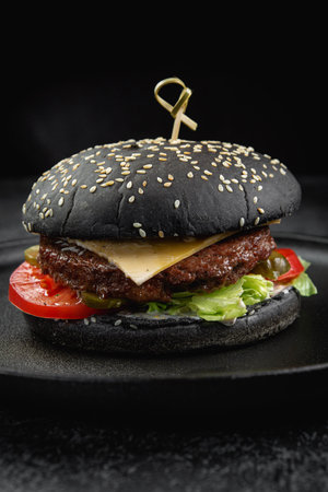 Black burger with a juicy meat patty, cheese, tomato, pickle on a dark stone tableの写真素材