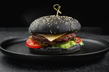 Black burger with a juicy meat patty, cheese, tomato, pickle on a dark stone tableの写真素材