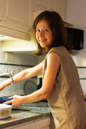 Smiling woman looking at camera while doing housework in kitchen, cleaning strawberriesの写真素材
