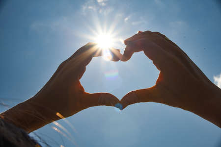 Heart shape universal sign for love and romance. Female hands in the shape of a heart on a background of bright sunの写真素材
