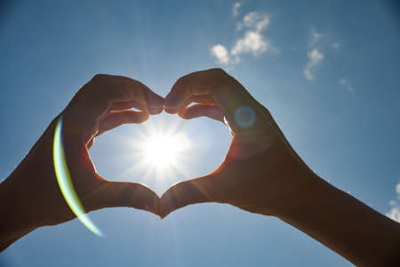 Heart shape universal sign for love and romance. Female hands in the shape of a heart on a background of bright sunの写真素材