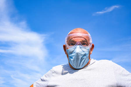 senior man wearing medical mask and goggles on a blue sky with clouds background. coronavirus concept. respiratory protectionの写真素材