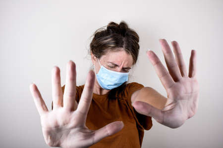 Woman is shocked in a medical mask and a brown T-shirt shows a stop signal with her hands isolated on white background. Woman encourages to stay at home.の写真素材