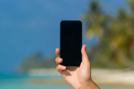 Woman hand showing a blank smart phone on the beach with the sea in the backgroundの写真素材