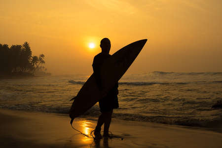 The male figure with a board for surf on a background of ocean waves. The setting sun paints the sky in bright orange yellow gold colorの写真素材
