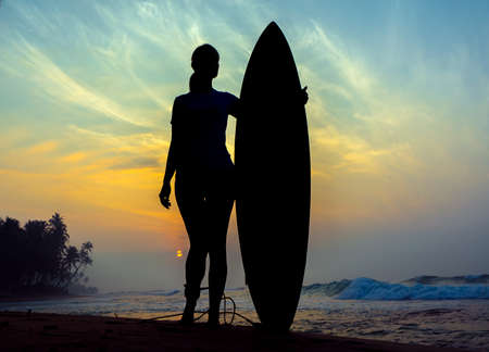 Surfer girl surfing looking at ocean beach sunset. Silhouette  woman looking at water with standing with surfboardの写真素材