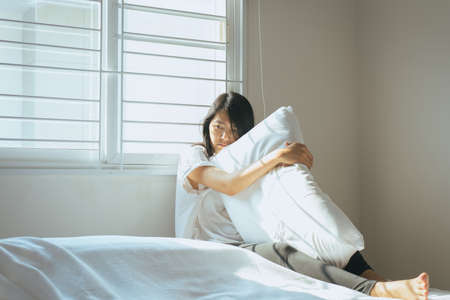 Asian woman have a thoughtful and hug the pillow on bed after wake up in the morning