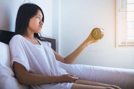 Young asian woman hates getting stressed waking up early,Female stretching her hand to ringing alarm to turn off alarm clock,Selective clockの写真素材