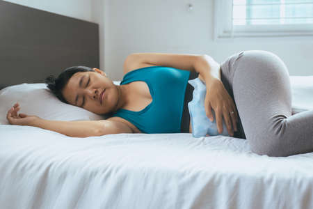 Woman holding hot water bag on her stomach in bedroom,Female lying in bedの写真素材