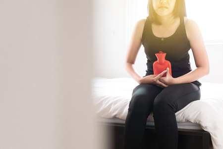 Female using hot water red bag or bottle on her stomach ache sitting on bedroomの写真素材