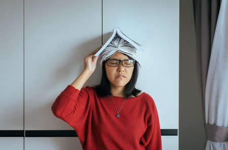 Asian woman holding a book on her head feelling serious and be tired at home