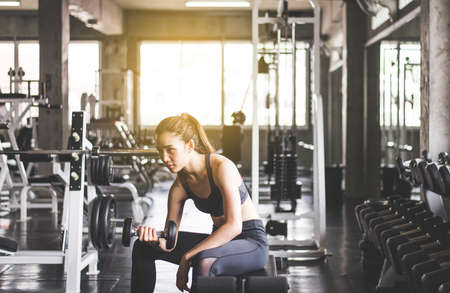 Sporty asian woman exercise with dumbbells sitting on bench,Female in sportswear does the exercises at gymの写真素材