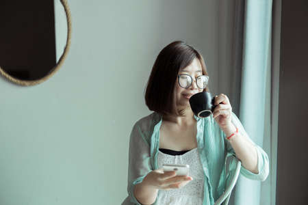 Portrait of beautiful asian woman is holding a cup of coffee and looking something on window at home in the morning,Happy and smilingの写真素材