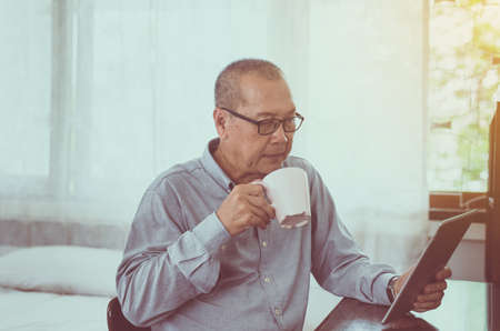 Asian senior man relaxing drinking coffee and using digital tablet at home in the morning
