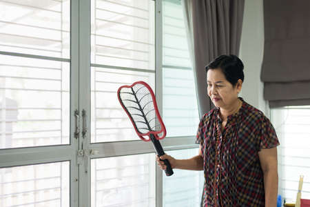 Middle aged woman using mosquito swatter at home,Female with mosquito electric net racket in livingの写真素材