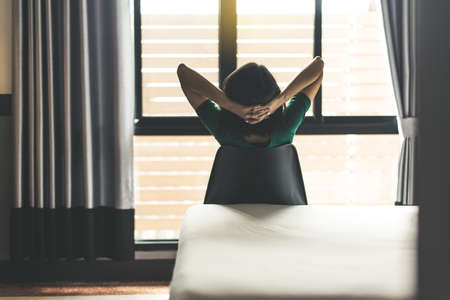 Back view of woman relaxing and sitting on chair with hands on her head and looking on window in bedroom at homeの写真素材