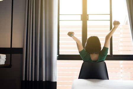 Back view of woman relaxing and sitting on chair with hands raise up and looking on window at house in the morningの写真素材