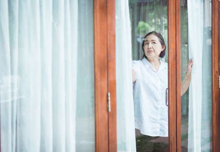 Senior healthy concept, Mature Asian elderly woman having migraine and looking something near window at homeの写真素材