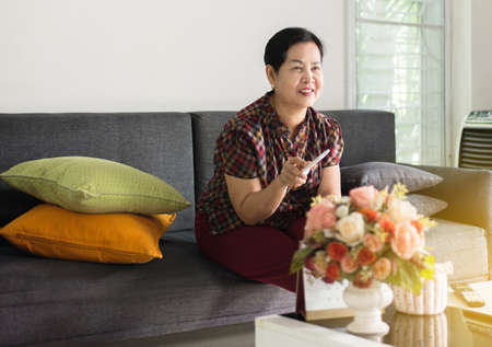 Happy elderly asian woman using remote and watching television on the couch in the living room at...