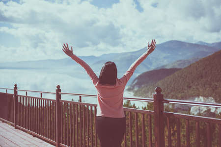 Happy and cheering woman stands with raised hands and looking beautiful amazing view at lake in the morningの写真素材