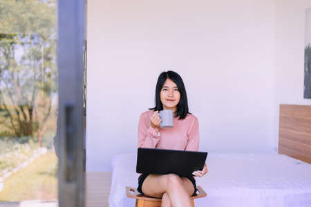 Happy asian woman drinking hot coffee after wake up and using laptop computer in bedroom, Relaxing timeの写真素材