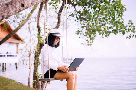 Beautiful asian woman sitting and using laptop computer on swing at sea, Happy and smiling, Relaxing timeの写真素材