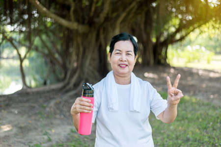 Senior asian woman showing two fingers and drinking water after break workout at park,Healthy...