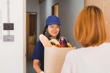 Delivery asian woman wearing face shield and holding vegetable with fresh fruits in crate send to customer at home,Delivers takeaway foodの写真素材