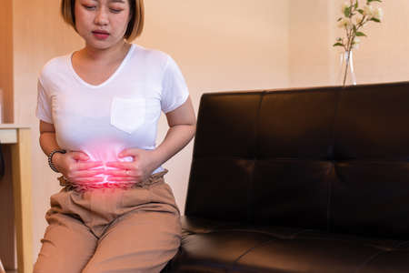 Asian woman having painful stomach ache,Female suffering from abdominal pain,Period cramps or premenstrual syndrome sitting on sofaの写真素材