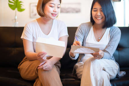 Happy two asian women writing greeting cards sitting on sofa at home for next holidayの写真素材