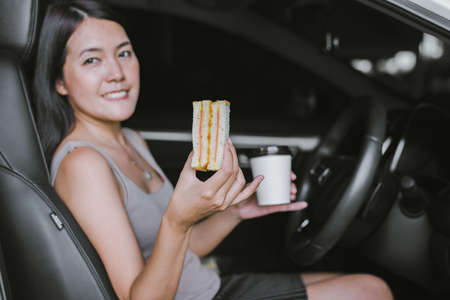Happy woman eating food and drinking coffee while driving to work in the early morningの写真素材