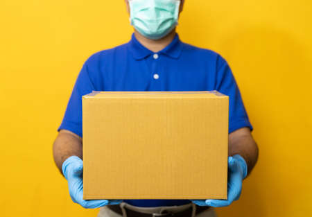 Delivery man blue uniform wearing rubber gloves and mask holding parcel cardboard box on yellow...
