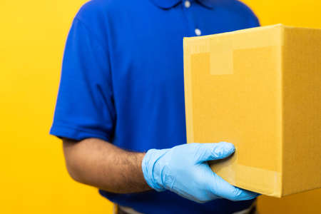 Delivery man blue uniform wearing rubber gloves holding parcel cardboard box on yellow background.