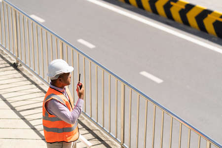 Asian civil engineer operate with radio to control working at construction. Worker wearing hard hat at highway concrete road site. Traffic report concept. Slow motion shot.の写真素材