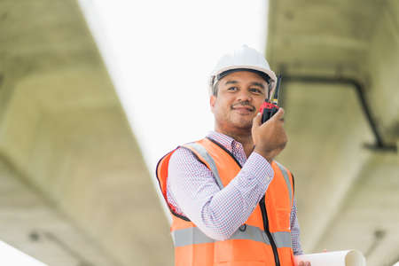 Asian civil engineer operate with radio talking to control working at construction. Worker wearing...