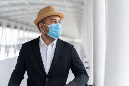 Young asian casual businessman wearing hat and protection mask standing on walkway airport terminal. Safety travel concept.の写真素材