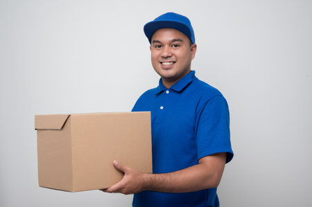 Young smiling asian delivery man in blue uniform holding box parcel cardboard on isolated white...