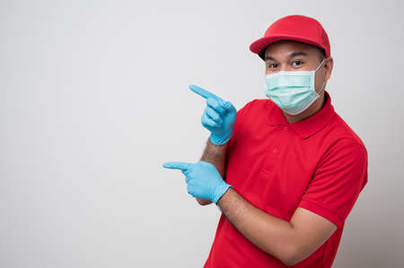 Young asian delivery man in red uniform wearing protection mask and medical rubber gloves giving the paper bag to customer on isolated white background. Safety deliver concept.の写真素材