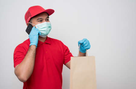 Young asian delivery man in red uniform wearing protection mask and medical rubber gloves using smartphone talking with customer to delivery parcel paper bag or food delivery.の写真素材
